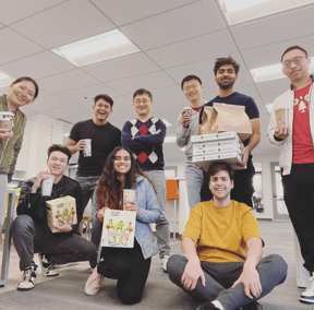 Nine people post for a photo as they hold pizza boxes and drinks for Hackathon event
